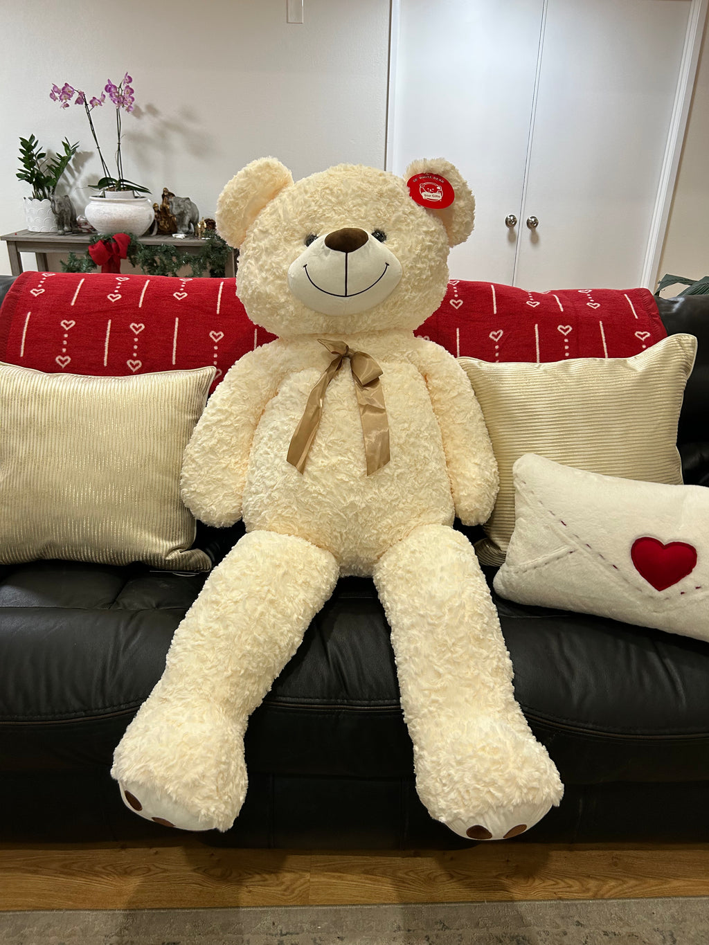Large white teddy bear with a bow tie sitting on a black couch with decorative pillows.