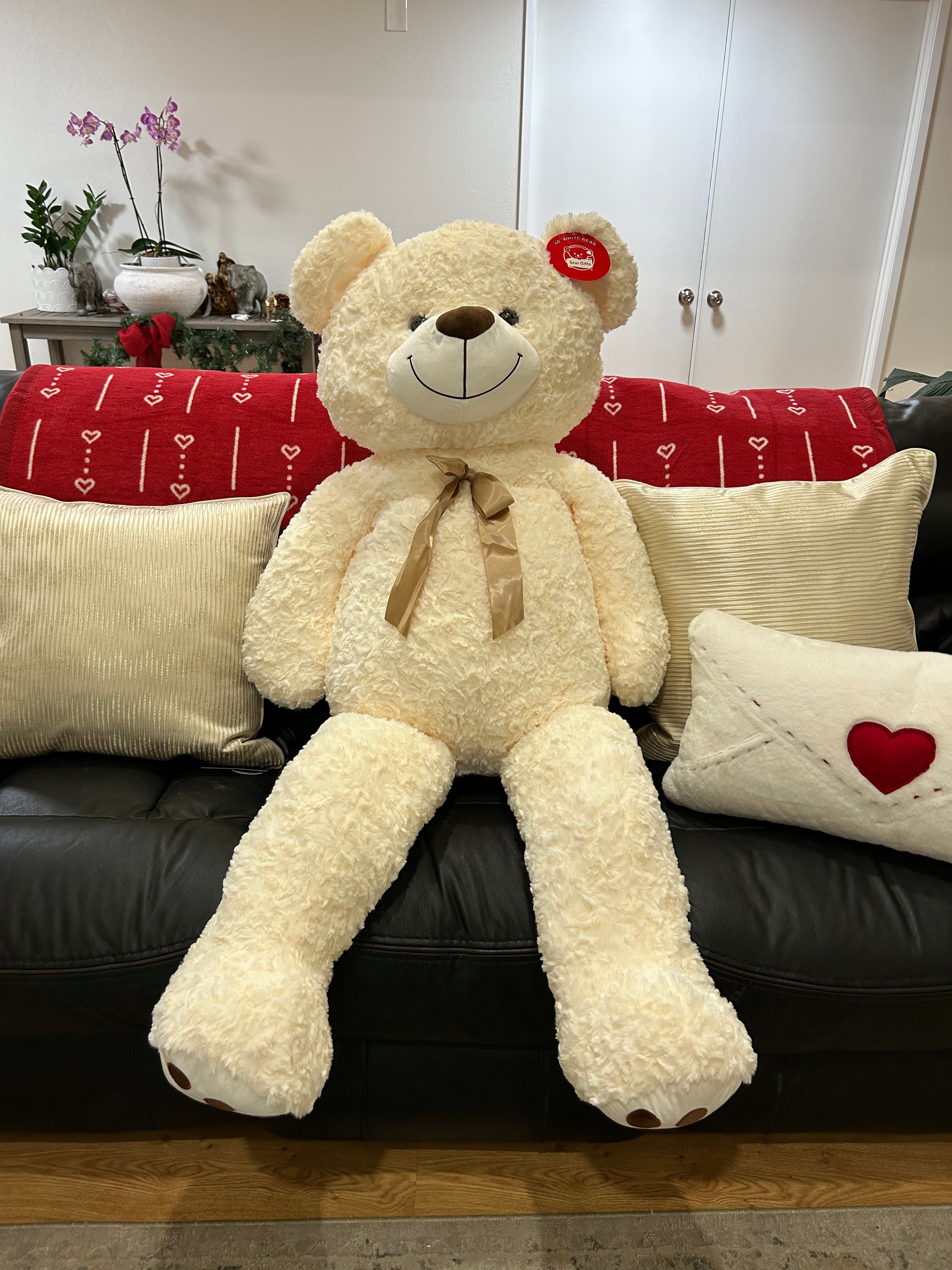 Large white teddy bear with a bow tie sitting on a black couch with decorative pillows.