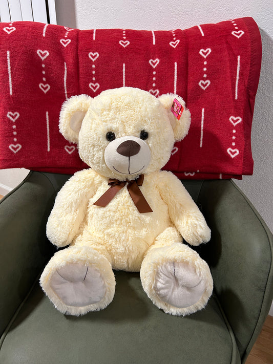 White teddy bear with a brown bow sitting on a green chair with a red blanket featuring heart patterns in the background.